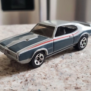May include: A die-cast model of a classic Pontiac GTO muscle car. The car is painted in a two-tone color scheme of gray and silver, with red and white accent stripes. The wheels are silver and black. The word "GTO" is visible on the side.