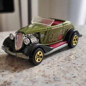 May include: A die-cast model of a vintage olive green roadster with black fenders and gold wheels. The car features a red interior and the text "Douglas" and "Orient" on the side. The car has a clear windshield and chrome detailing.