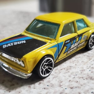 May include: A yellow Datsun 510 toy car with black and blue racing stripes on the hood. The car has black wheels with silver rims and the text "77 Racing" on the side.
