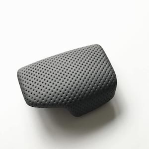 May include: A dark grey, rectangular automotive armrest cover featuring a uniform, perforated texture across its entire surface. The piece has rounded corners and a contoured shape designed to fit a specific vehicle console component.