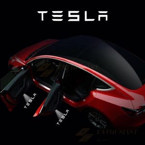 May include: A red Tesla car with the doors open, displaying the word "TESLA" in white. The car has a black roof and the word "TESLA" is also projected onto the ground from the open doors. The word "ENTHUSIAST" is also visible.