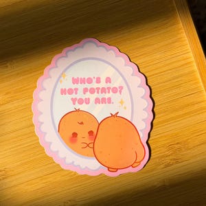 May include: A die-cut sticker featuring a scalloped pink border and a mirror illustration. The text reads, "WHO'S A HOT POTATO? YOU ARE." A cartoon potato stands with its back to the viewer, looking at its reflection in the mirror.