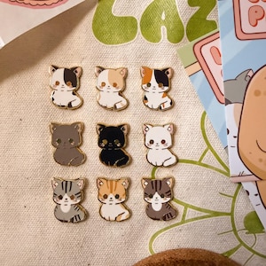 Könnte beinhalten: Neun kleine Emaille-Anstecknadeln in Form von sitzenden Katzen in verschiedenen Farben und Mustern, darunter einfarbig schwarz, weiß, grau sowie mehrfarbige Glückskatzen- oder getigerte Designs. Jede Nadel hat goldfarbene Metallränder.
