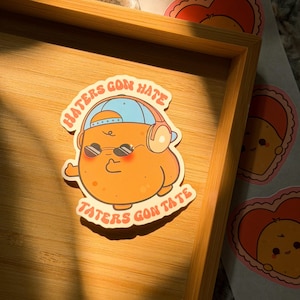 Peut inclure: Un autocollant découpé représentant un personnage de pomme de terre de dessin animé portant une casquette de baseball bleue, un casque audio et des lunettes de soleil. La pomme de terre sourit et fait un pouce levé. Le texte indique "HATERS GON HATE" et "TATERS GON TATE" en orange.