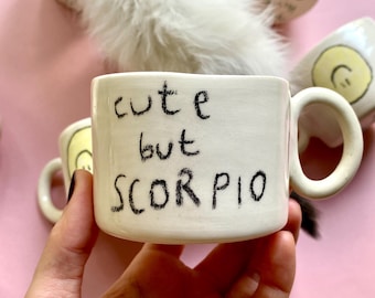 May include: White ceramic mug with black handwritten text that reads "cute but scorpio".