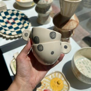 Puede incluir: Dos tazas de t&eacute; de cer&aacute;mica blanquecina con lunares negros y asas redondas. Las tazas est&aacute;n apiladas, y en el fondo se ven otras piezas de cer&aacute;mica con varios dise&ntilde;os.