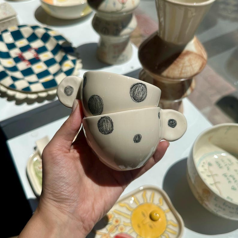 Puede incluir: Dos tazas de t&eacute; de cer&aacute;mica blanquecina con lunares negros y asas redondas. Las tazas est&aacute;n apiladas, y en el fondo se ven otras piezas de cer&aacute;mica con varios dise&ntilde;os.