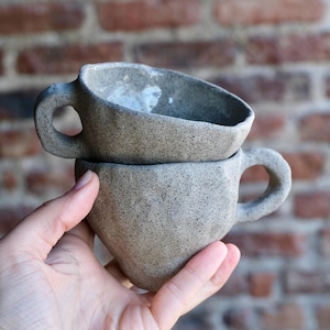 May include: Two gray ceramic mugs stacked on top of each other. Each mug has a rounded handle and a textured surface. The mugs are held in a hand, with a brick wall in the background.