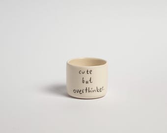 May include: A white ceramic mug with the text "cute but overthinker" written in black.