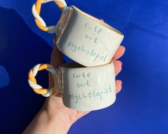 May include: Two white ceramic mugs with twisted handles. The mugs have the text "cute but psychologist" written on them in green.