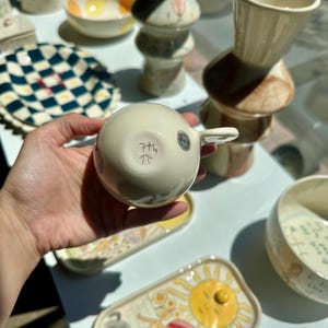 Puede incluir: Una taza de t&eacute; de cer&aacute;mica color crema con asa, con un dise&ntilde;o escrito a mano en la parte inferior. La taza forma parte de una colecci&oacute;n de cer&aacute;mica hecha a mano, que incluye platos y jarrones decorativos. La taza de t&eacute; est&aacute; en una mano.