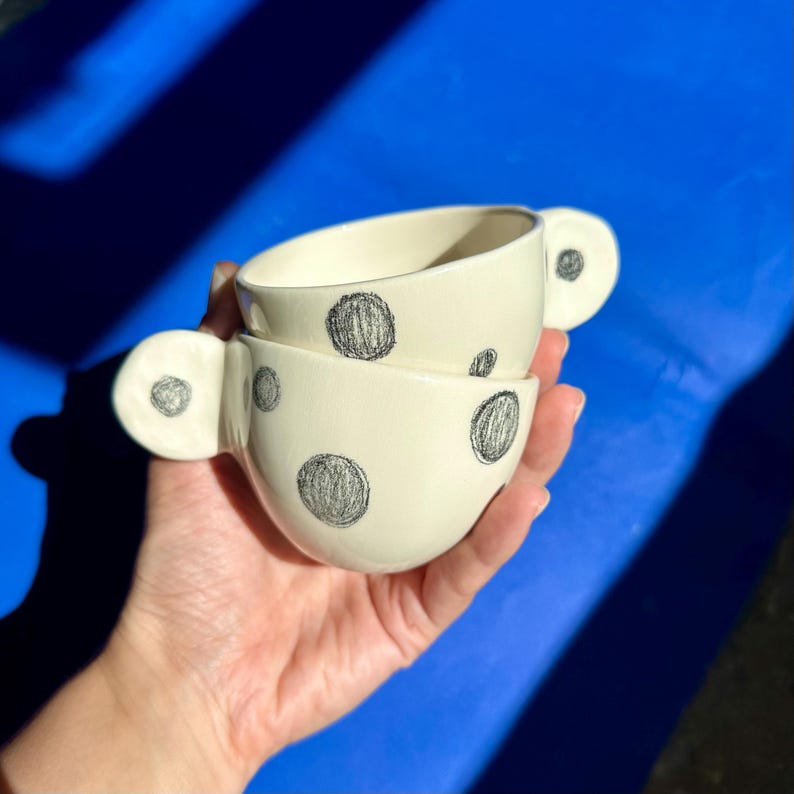 Puede incluir: Dos tazas de cer&aacute;mica blanquecina con grandes lunares negros dibujados a mano. Cada taza tiene un asa redondeada y est&aacute;n apiladas una encima de la otra. Las tazas se sostienen en una mano sobre un fondo azul.