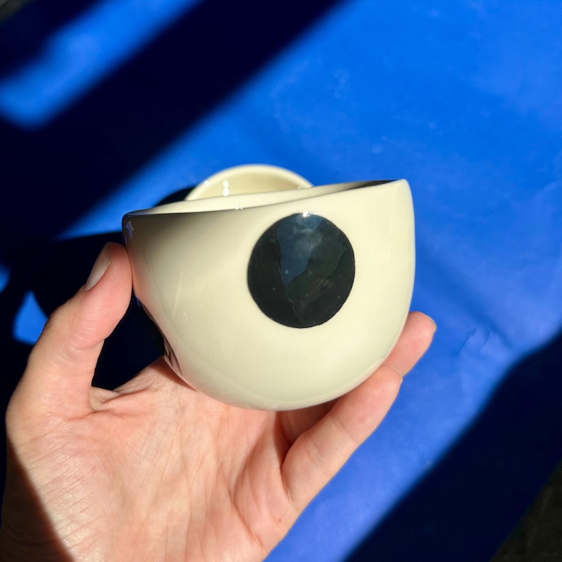 Może przedstawiać: Kremowa ceramiczna miska z dużym czarnym okręgiem z boku. Miska ma organiczny kształt i mały uchwyt. Miska jest trzymana w dłoni na niebieskim tle.