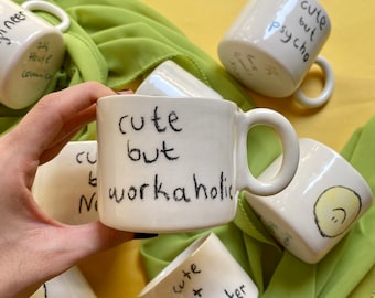 May include: A collection of white ceramic mugs with black handwritten text. One mug reads "cute but workaholic," another "cute but psycho." Other mugs have similar phrases and a smiley face. The mugs are on a yellow surface with green fabric.