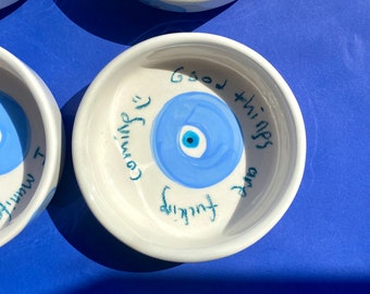 May include: White ceramic dish with a blue evil eye design and the text "Good things are coming" written around the eye.
