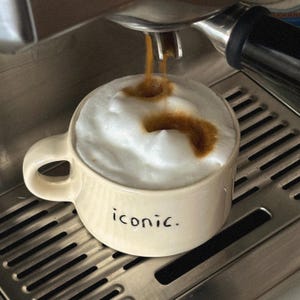 May include: A beige ceramic mug with the word "iconic" in black lettering is filled with a cappuccino. The coffee is being poured from an espresso machine, creating a foamy top.