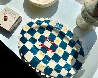 May include: A ceramic serving platter with a blue and white checkered pattern and a scalloped edge. The words "Ciao Amore" are written in red. Small yellow lemon illustrations are scattered across the surface. Other ceramic bowls and containers are also visible.