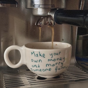 Pode incluir: Uma caneca de café de cerâmica branca com uma alça grande está posicionada sob uma máquina de café enquanto o café expresso é derramado. A caneca tem o texto manuscrito "Make your own money and marry someone funny."