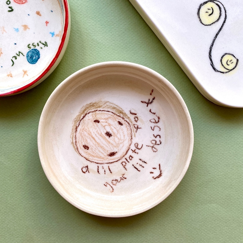 May include: A white ceramic plate with brown hand-drawn illustrations of a smiling face and the text "your little plate for dessert".