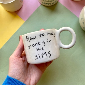 May include: A white ceramic mug with a rounded handle. The mug has the handwritten text "How to make money in the SIMS" in black ink. The mug is held in a hand against a colorful background.