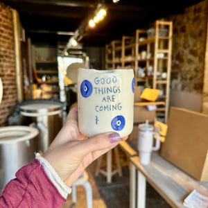 May include: White ceramic mug with blue evil eye charms and the text "Good things are coming".