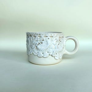May include: A white ceramic mug with a textured lace design around the top. The mug has a rounded handle and a smooth, off-white finish. The lace detail adds a delicate, handcrafted aesthetic to this drinkware.