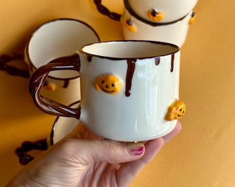 May include: Cream-colored ceramic mugs with brown dripped accents and handles. Each mug features orange pumpkin embellishments. The mugs are stacked and arranged on a yellow background, creating a festive Halloween theme.