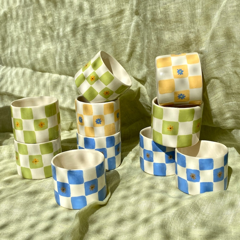 May include: A set of eight ceramic cups with a checkered pattern in blue, green, and yellow with small flower accents. The cups are stacked and arranged on a green fabric surface.