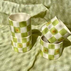 May include: Four ceramic cups with a green and white checkered pattern and small yellow flowers. The cups are stacked on top of each other.