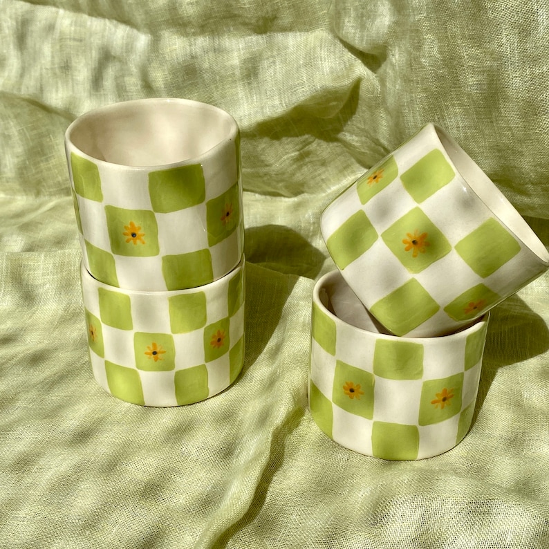 May include: Four ceramic cups with a green and white checkered pattern and small yellow flowers. The cups are stacked on top of each other.