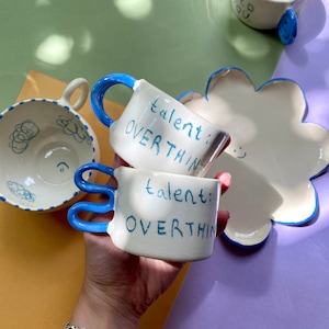 May include: Two white ceramic mugs with blue handles. The mugs have the text "talent: overthink" written on them in blue.