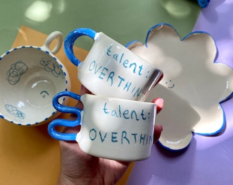 May include: Two white ceramic mugs with blue handles. The mugs have the text "talent: overthink" written on them in blue.