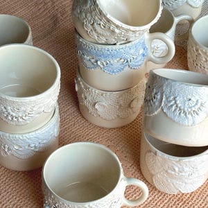 Peut inclure: Une collection de tasses en céramique avec une base crème et des détails complexes en forme de dentelle. Certaines tasses présentent un accent de dentelle bleu clair. Les tasses ont une anse et sont empilées sur une surface rose.