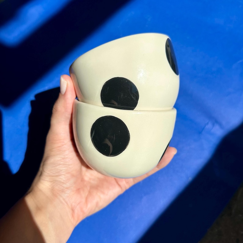 Może przedstawiać: Dwie kremowe ceramiczne miski ułożone jedna na drugiej, każda z dużą czarną kropką. Miski są trzymane w dłoni na niebieskim tle. Miski mają zaokrąglony kształt.