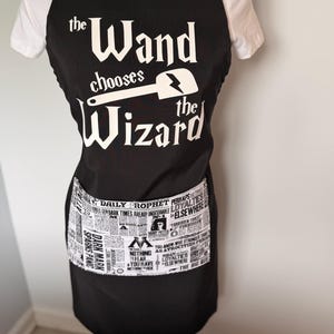 May include: A black apron featuring white text that reads "the Wand chooses the Wizard" with a spatula graphic. The apron includes a front pocket with a black and white newspaper print pattern featuring headlines and text from the Daily Prophet.
