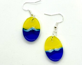 May include: A pair of oval-shaped earrings with a blue and yellow design. The earrings are made of glass and have silver hooks.