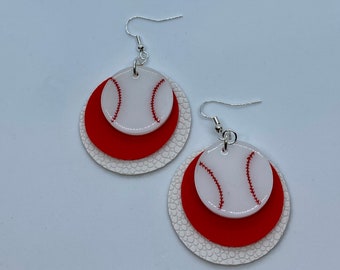 Könnte beinhalten: Ohrringe im Baseball-Design. Sie zeigen ein weißes Baseball-Design mit roten Nähten, das über einem roten Kreis und einem strukturierten weißen Kreis angeordnet ist. Die Ohrringe haben silberfarbene Haken.