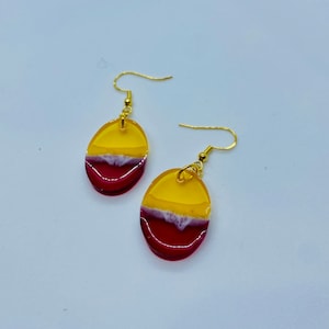 May include: A pair of oval-shaped earrings with a yellow, white, and red resin design. The earrings have gold-colored hooks.