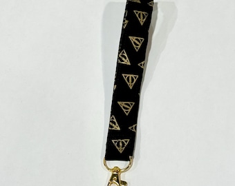 May include: Black lanyard with gold-colored Deathly Hallows symbols. The lanyard has a gold-colored clasp at the bottom. The Deathly Hallows symbol is a triangle, a circle, and a vertical line.