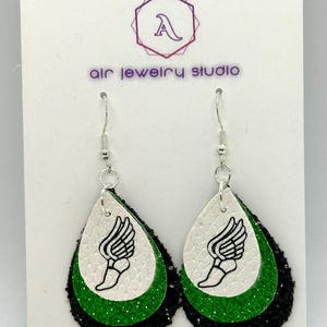 May include: A pair of dangle earrings with a green, black, and white layered design. The white layer features a black and white graphic of a winged foot. The earrings are on a white card with the text "Air Jewelry Studio" printed on it.