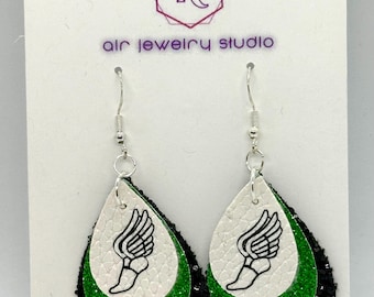 May include: A pair of dangle earrings with a green, black, and white layered design. The white layer features a black and white graphic of a winged foot. The earrings are on a white card with the text "Air Jewelry Studio" printed on it.