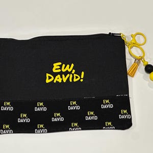 May include: Black canvas pouch with the text "EW, DAVID!" in yellow. The bottom of the pouch has a repeating pattern of "EW, DAVID" in white and yellow. A yellow scissor-shaped keychain with a tassel and beaded accents is attached.