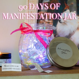May include: A glass jar with a pink ribbon and string lights inside. The jar is labeled "Manifestation Station". There is a small candle and a piece of paper with the text "Money comes to me effortlessly and easily." on a wooden surface.