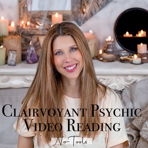 May include: A woman with long brown hair smiles at the camera. She is wearing a light beige shirt and sitting on a couch. The text "Clairvoyant Psychic Video Reading No Tools" is displayed in black text on the front of her shirt. The background features a fireplace mantel with candles and other decorative items.