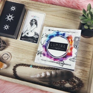 May include: A wooden tray with a variety of items, including a deck of tarot cards, a book titled "LUMIWA TAROT", a small Buddha statue, a crystal, a beaded necklace, a feather, and a small potted succulent.