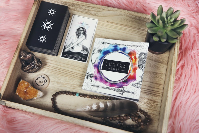 May include: A wooden tray with a variety of items, including a deck of tarot cards, a book titled "LUMIWA TAROT", a small Buddha statue, a crystal, a beaded necklace, a feather, and a small potted succulent.