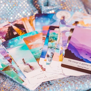 May include: A deck of oracle cards with a pastel color scheme and illustrations of celestial bodies, mountains, and water. The top card reads "The energy is gaining momentum. Waxing Moon."