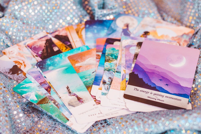 May include: A deck of oracle cards with a pastel color scheme and illustrations of celestial bodies, mountains, and water. The top card reads "The energy is gaining momentum. Waxing Moon."