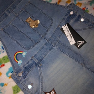 Puede incluir: Mono vaquero azul claro con parches decorativos, incluyendo un oso de peluche, un arcoíris y un búho. El mono tiene botones plateados y una etiqueta que dice "SMART DENIM" y "Jordache".