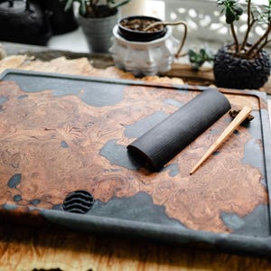 May include: A rectangular tea tray with a dark gray border and a wood grain pattern in shades of brown and gray. A black tea tool and a wooden tea pick rest on the tray. The tray has a small, round drainage hole.